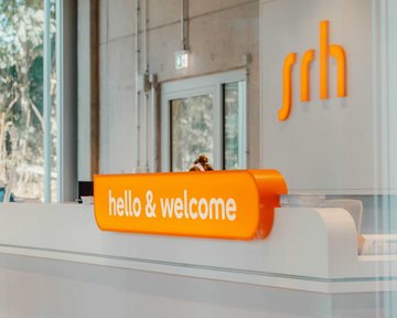 Empfangstresen mit der Aufschrift "hello & welcome" und dem Logo "srh" in orange. Helle, moderne Umgebung mit Fenstern.