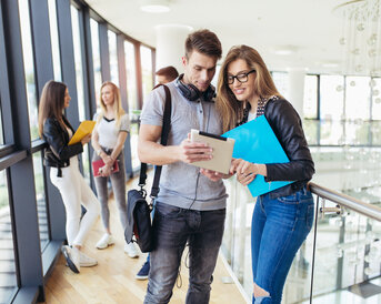 Zwei Studierende schauen auf ein Tablet, während sie in einem modernen Gebäude stehen. Im Hintergrund sind weitere Studierende zu sehen.