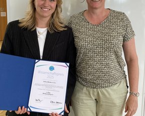 Zwei Frauen stehen nebeneinander, eine hält ein Zertifikat für den Wissenschaftspreis 2025 in der Hand. Beide lächeln in die Kamera.