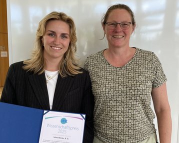 Zwei Frauen stehen nebeneinander, eine hält ein Zertifikat für den Wissenschaftspreis 2025 in der Hand. Beide lächeln in die Kamera.