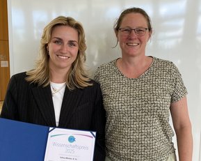Zwei Frauen stehen nebeneinander, eine hält ein Zertifikat für den Wissenschaftspreis 2025 in der Hand. Beide lächeln in die Kamera.