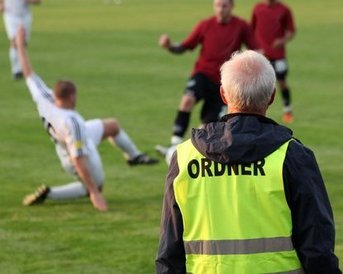 Ein Sicherheitsmitarbeiter in einer gelben Weste beobachtet ein Fußballspiel, während ein Spieler auf dem Rasen gefallen ist.