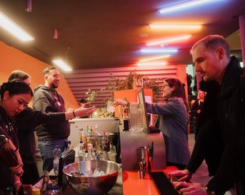 A group of people at a bar, preparing and serving drinks, surrounded by colorful neon lights.