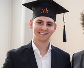 Ein junger Mann in einem Anzug mit einem Abschlusshut steht neben einem älteren Mann in einem Anzug mit Brille.