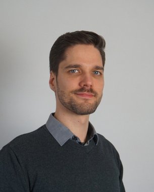 Ein Mann mit kurzen, braunen Haaren und einem Bart trägt einen grauen Pullover und ein hellblaues Hemd. Er blickt zur Seite.