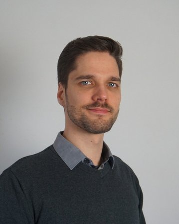 Ein Mann mit kurzen, braunen Haaren und einem Bart trägt einen grauen Pullover und ein hellblaues Hemd. Er blickt zur Seite.