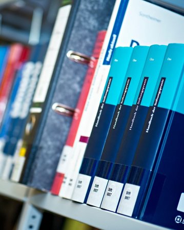 Bookshelf with various colored spines, including several volumes in blue and black. Some books are placed in folders, while others stand upright. The titles are partially visible but not fully legible.