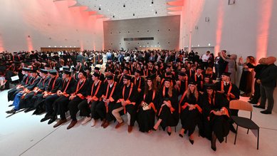 Graduates in black gowns and red sashes are seated in a festive hall during a graduation ceremony.
