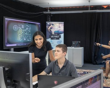 A woman is showing a man something on the computer, while in the background, other people are working with VR technology.