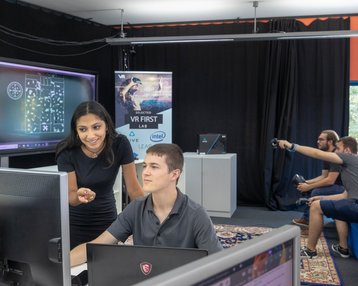 Eine Frau zeigt einem Mann am Computer etwas, während im Hintergrund andere Personen an VR-Technologie arbeiten.
