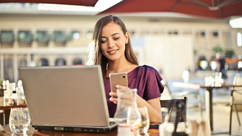 Eine Frau sitzt an einem Tisch im Freien, lächelt und schaut auf ihr Smartphone, während ein Laptop vor ihr steht. Im Hintergrund sind Tische und Stühle eines Cafés sichtbar.