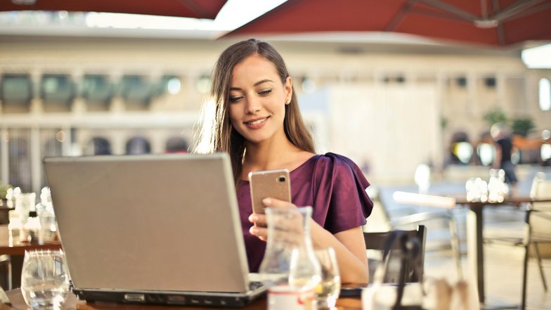 Eine Frau sitzt an einem Tisch im Freien, lächelt und schaut auf ihr Smartphone, während ein Laptop vor ihr steht. Im Hintergrund sind Tische und Stühle eines Cafés sichtbar.