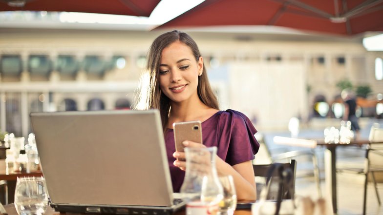 Eine Frau sitzt an einem Tisch im Freien, lächelt und schaut auf ihr Smartphone, während ein Laptop vor ihr steht. Im Hintergrund sind Tische und Stühle eines Cafés sichtbar.