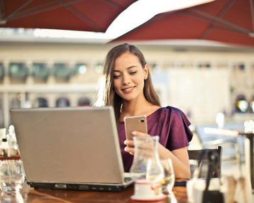 Eine Frau sitzt an einem Tisch im Freien, lächelt und schaut auf ihr Smartphone, während ein Laptop vor ihr steht. Im Hintergrund sind Tische und Stühle eines Cafés sichtbar.