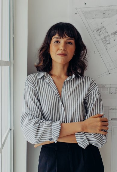 A woman with curly hair stands smiling in front of an architectural plan on the wall. She is wearing a striped shirt and has her arms crossed. Daylight streams in through a window beside her.