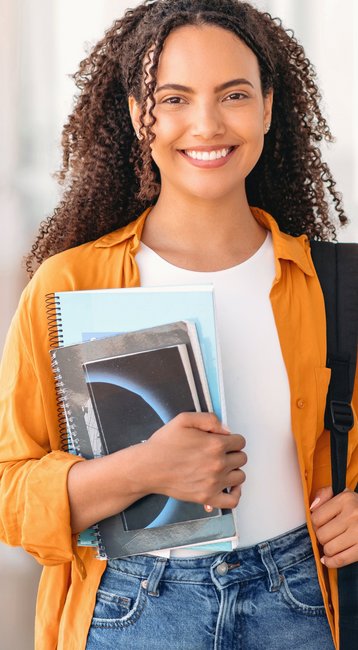 Eine junge Frau mit lockigem Haar trägt eine orangefarbene Jacke und hält mehrere Bücher in der Hand. Sie steht in einem modernen Flur und lächelt, während sie eine schwarze Tasche über der Schulter trägt.