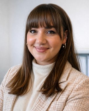 Eine lächelnde Frau mit langen, braunen Haaren trägt einen hellen Blazer und einen Rollkragenpullover. Sie steht vor einer neutralen Wand.
