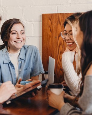 Four people are sitting at a table in a café. A man is using a tablet while the other three women are smiling and chatting with each other. Drinks are placed on the table, and the atmosphere feels relaxed.