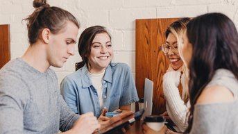 Four people are sitting at a table in a café. A man is using a tablet while the other three women are smiling and chatting with each other. Drinks are placed on the table, and the atmosphere feels relaxed.