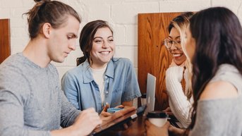 Vier Personen sitzen an einem Tisch in einem Café. Ein Mann nutzt ein Tablet, während die anderen drei Frauen lächeln und miteinander sprechen. Getränke sind auf dem Tisch platziert, und die Atmosphäre wirkt entspannt.