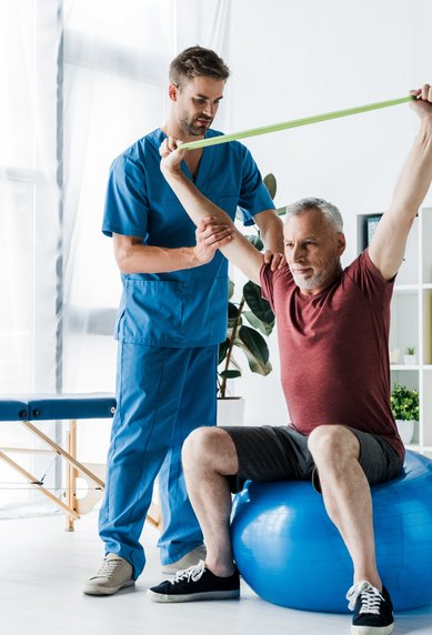 Ein Physiotherapeut hilft einem älteren Mann, der auf einem Gymnastikball sitzt, mit einem Widerstandsband beim Training.