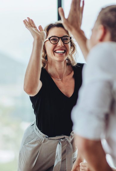 A group of people in a modern office, with a woman wearing glasses enthusiastically gesturing with her hands.