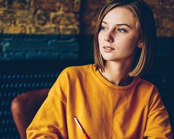 Eine junge Frau mit schulterlangen Haaren trägt ein oranges Sweatshirt und schaut nachdenklich, während sie einen Stift in der Hand hält.