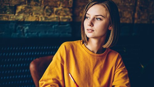 Eine junge Frau mit schulterlangen Haaren trägt ein oranges Sweatshirt und schaut nachdenklich, während sie einen Stift in der Hand hält.