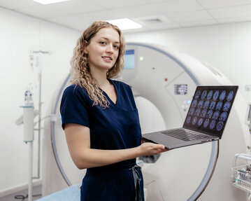 Eine medizinische Fachkraft hält einen Laptop mit Gehirnscans in einem modernen CT-Raum. Im Hintergrund ist das CT-Gerät sichtbar.