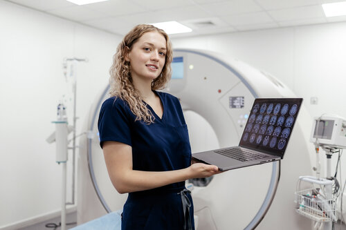 Eine medizinische Fachkraft hält einen Laptop mit Gehirnscans in einem modernen CT-Raum. Im Hintergrund ist das CT-Gerät sichtbar.