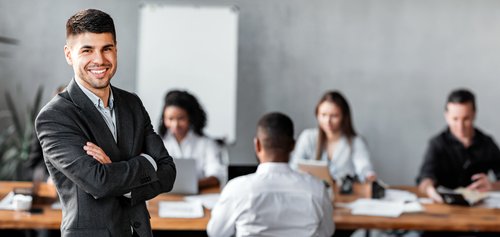 Ein lächelnder Mann in einem Anzug steht vor einer Gruppe von Menschen, die an einem Tisch arbeiten. Im Hintergrund sind Notizen und Laptops sichtbar.