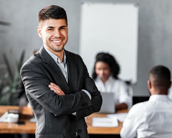 A smiling man in a suit stands in front of a group of people working at a table. In the background, notes and laptops are visible.