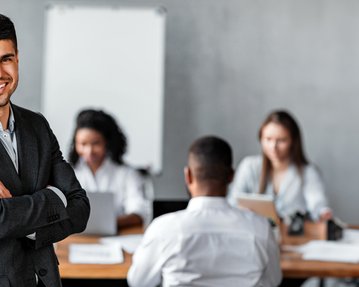 A smiling man in a suit stands in front of a group of people working at a table. In the background, notes and laptops are visible.