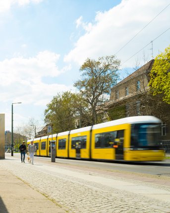 A yellow streetcar glides along a wall while two people stroll on the sidewalk. Bright clouds fill the sky.