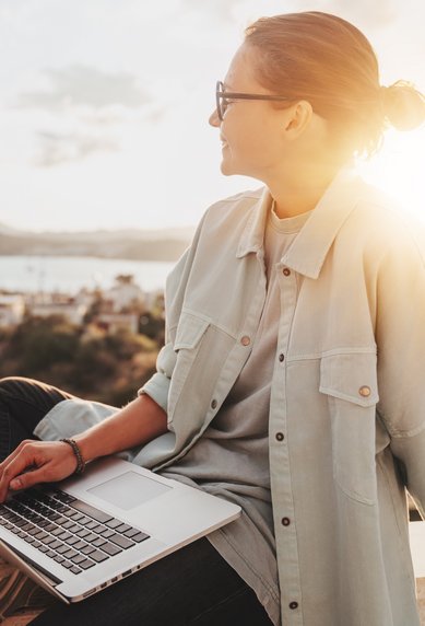 Eine Person sitzt im Freien mit einem Laptop auf dem Schoß und blickt auf eine Stadtlandschaft im Hintergrund, während die Sonne untergeht.