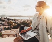 Eine Person sitzt im Freien mit einem Laptop auf dem Schoß und blickt auf eine Stadtlandschaft im Hintergrund, während die Sonne untergeht.