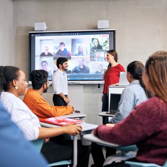 A group of students is sitting in a classroom, looking at a screen displaying a video conference with several participants. Two people are standing at the front and talking to each other.