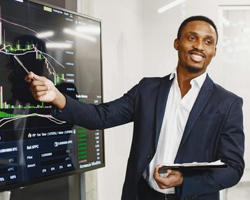Ein Mann in einem Anzug zeigt auf ein Finanzdiagramm auf einem Bildschirm und erklärt die Daten. Er hält ein Notizbuch in der Hand und lächelt, während er seine Präsentation durchführt.