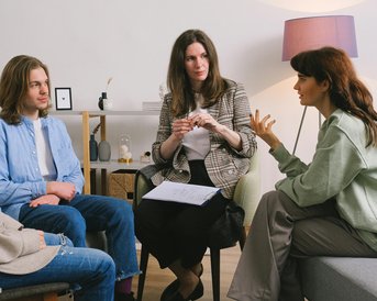Eine Gruppe von vier Personen sitzt in einem hellen Raum. Eine Frau spricht, während die anderen aufmerksam zuhören. Es gibt eine entspannte Atmosphäre mit modernen Möbeln und einer Lampe im Hintergrund.