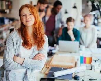 Eine lächelnde Frau mit roten Haaren steht im Vordergrund, während mehrere Personen im Hintergrund an einem Tisch arbeiten.