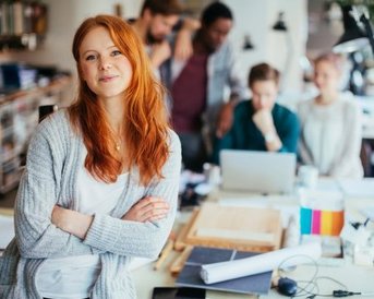 A smiling woman with red hair stands in the foreground, while several people in the background are working at a table.