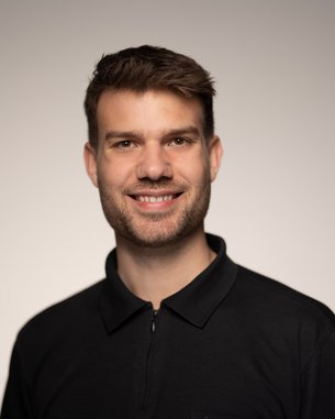 Ein junger Mann mit kurzen, braunen Haaren und einem freundlichen Lächeln trägt ein schwarzes Polohemd. Der Hintergrund ist neutral und hell.