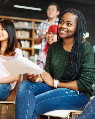 Eine Gruppe von jungen Menschen sitzt in einem Raum mit Bücherregalen, aufmerksam und lächelnd. Einige halten Papiere in den Händen, während sie auf eine Präsentation oder Diskussion warten.