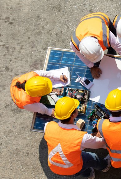 Six construction workers in safety vests and helmets are discussing plans and working at a table with technical equipment and blueprints.