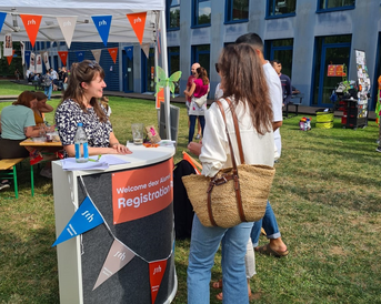A person is standing at a registration table decorated with colorful bunting. In the background, there are more people and booths visible. The scene takes place outdoors on a lawn, surrounded by buildings.