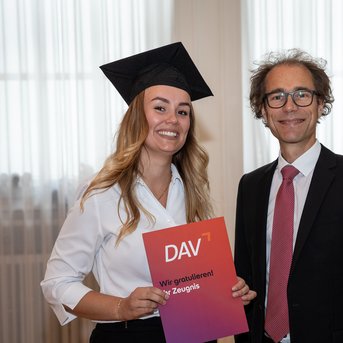 Eine graduierte Person in schwarzer Mütze hält ein Zertifikat mit der Aufschrift "DAV". Neben ihr steht ein Mann in Anzug.