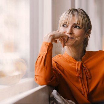 A smiling woman with blonde hair sits by the window, looking thoughtfully. She is wearing an orange blouse and has one hand resting on her chin. In the background, blurred lights can be seen.