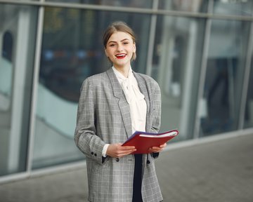 Eine junge Frau steht im Freien und hält einen roten Ordner. Sie trägt einen grauen Blazer und eine weiße Bluse. Ihr Haar ist locker frisiert, und sie lächelt freundlich in die Kamera.