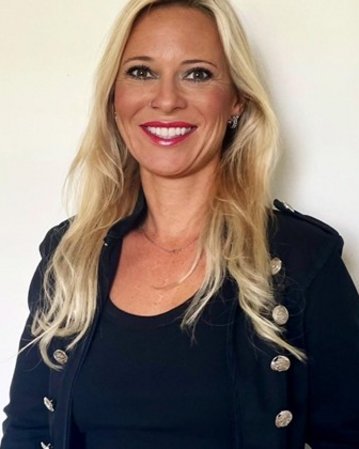 A woman with long blonde hair is wearing a black jacket with gold buttons and is smiling warmly at the camera.