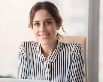 A woman with long brown hair is sitting at a desk and smiling. She is wearing a striped shirt and has a laptop in front of her. In the background, there are blurred plants and windows visible.